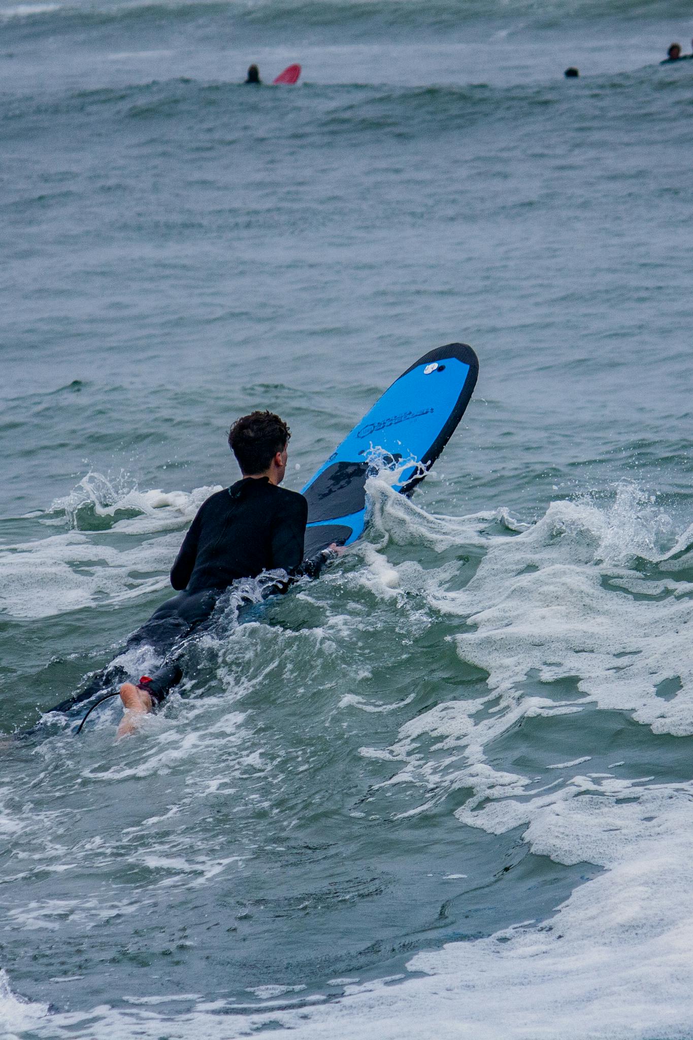 A surfer in a wetsuit paddles through ocean waves, positioning for the next surf ride on an overcast day.