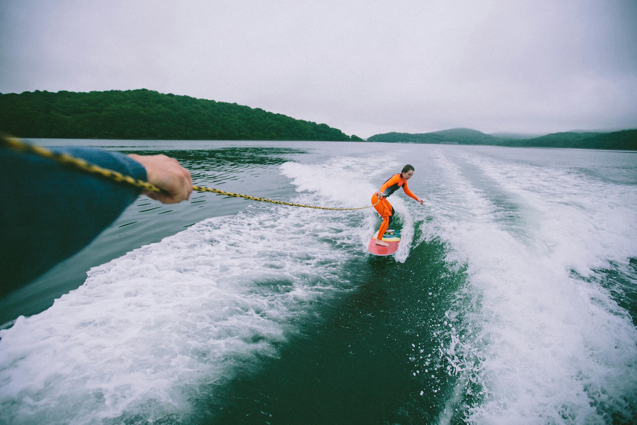Woman wakesurfing on a lake, wearing a bright wetsuit, balancing on the board with a rope in hand.